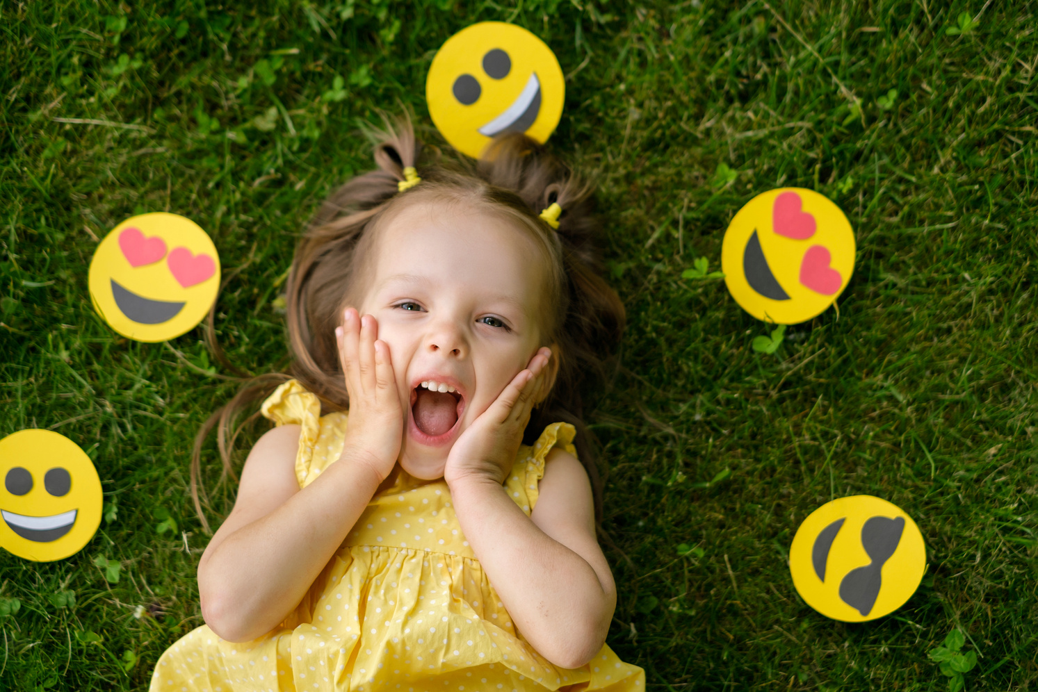 A funny little girl surrounded by a variety of paper emoticons looks up in surprise with her mouth open in delight. Emotional little kid celebrates world smile day